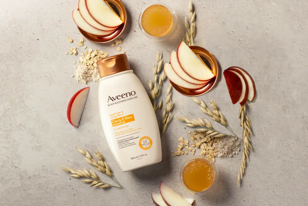 A bottle of Aveeno® Clarity & Shine Shampoo displayed with apple slices, apple cider vinegar and oats 