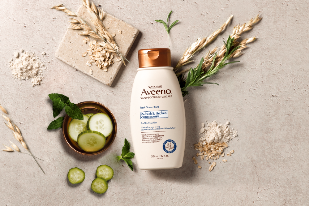 A bottle of Aveeno® Refresh & Thicken Conditioner displayed with cucumber slices, herbs and oats