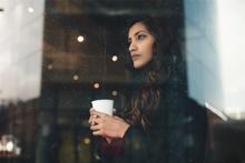 A woman with seasonal affective disorder holds a mug and looks out a window.