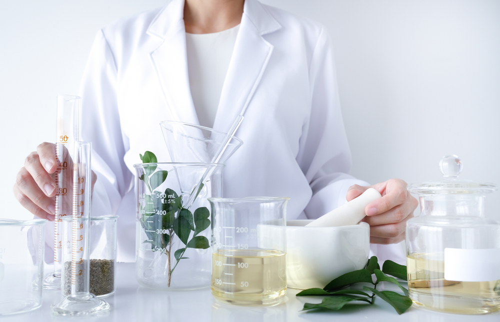 A clinician in a white coat works at a table of beakers filled with natural skincare ingredients.