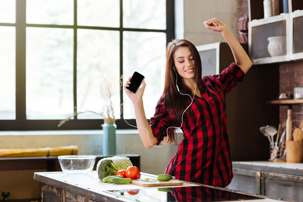 Woman in red shirt with eyes closed listening to music, dancing in kitchen with phone and headphone.